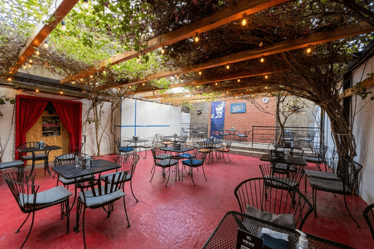 Patio de Ocho y Medio Café en Mexicali: pérgola de madera cubierta de enredadera con foquitos, mesas de herrería negra y piso rojo