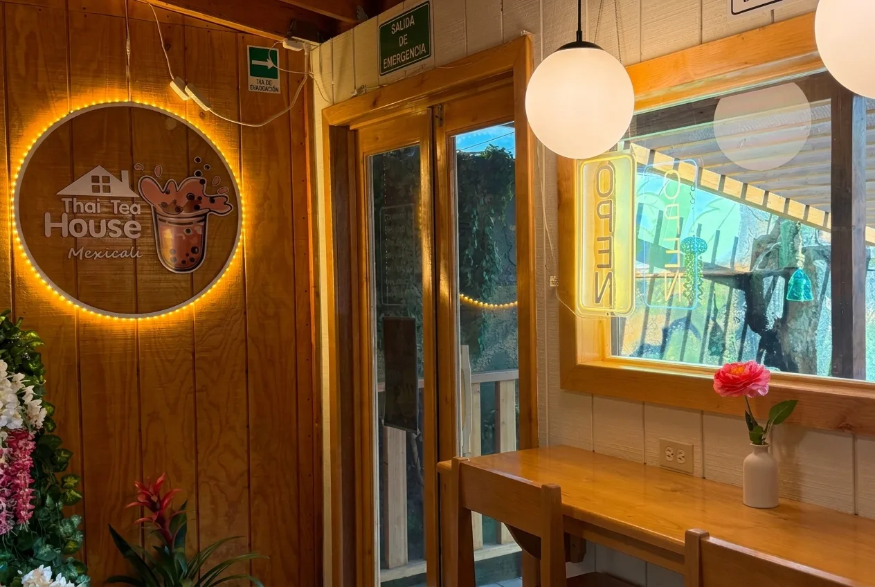 Interior de Thai Tea House Mexicali: pared de madera con su logo redondo iluminado, puerta de cristal, ventana con letrero OPEN y barra de madera con una flor